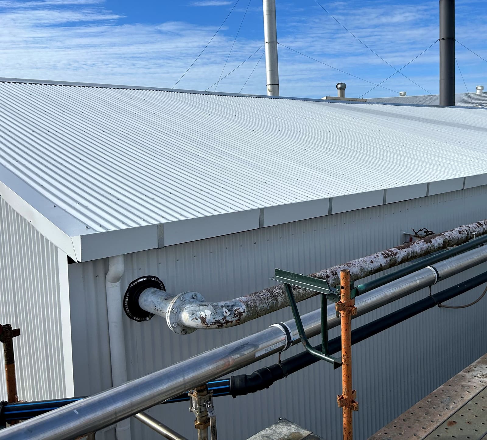 New pale corrugated roof sheeting and matching wall cladding on a commercial building, with two flue stacks and scaffold alongside.