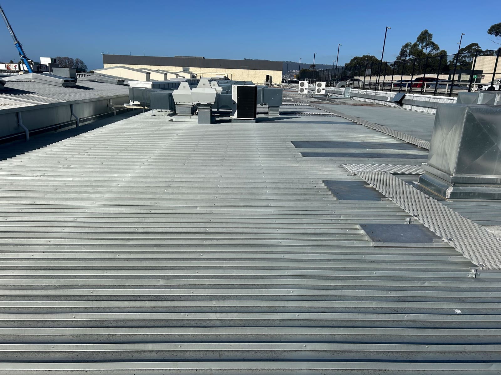 A wide commercial metal roof with ducting, plant units and walkway mesh, and a crane on the skyline.